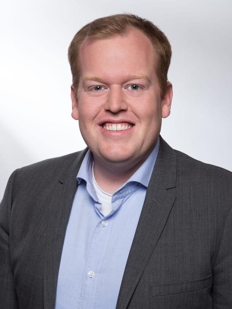 Porträt eines lächelnden Mannes in einem grauen Anzug mit hellblauer Hemd unter neutralem Hintergrund.