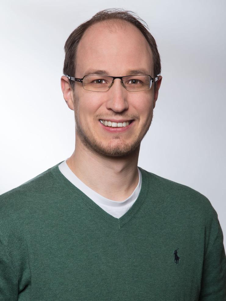 Portrait eines Mannes mit Brille, grünem Pullover und weißem T-Shirt, lächelnd vor neutralem Hintergrund.
