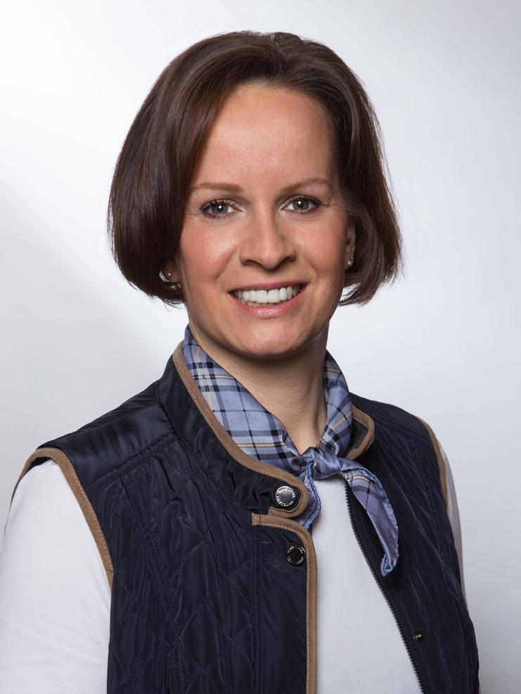 Lächelnde Frau mit schulterlangen Haaren, gekleidet in eine gestickte Weste und kariertem Halstuch, vor hellem Hintergrund.