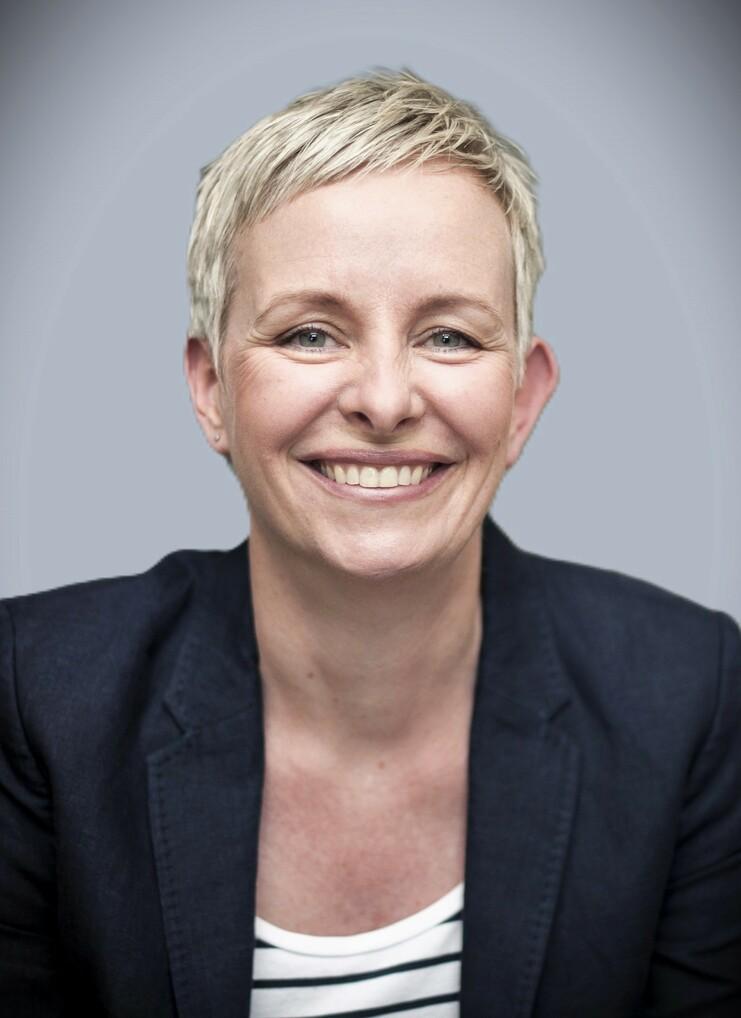 Porträt einer lächelnden Frau mit kurzer, heller Frisur, die einen blauen Blazer und ein gestreiftes Oberteil trägt.