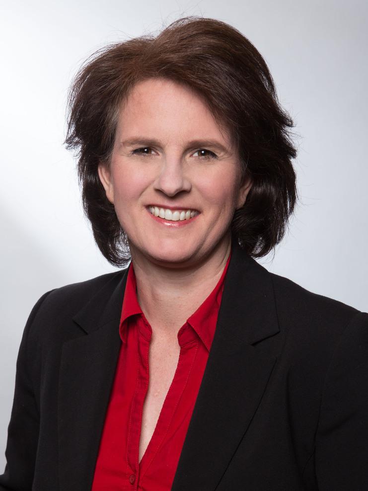 Porträt einer lächelnden Frau mit braunem Haar, bluse in Rot und schwarzem Blazer vor einem hellen Hintergrund.