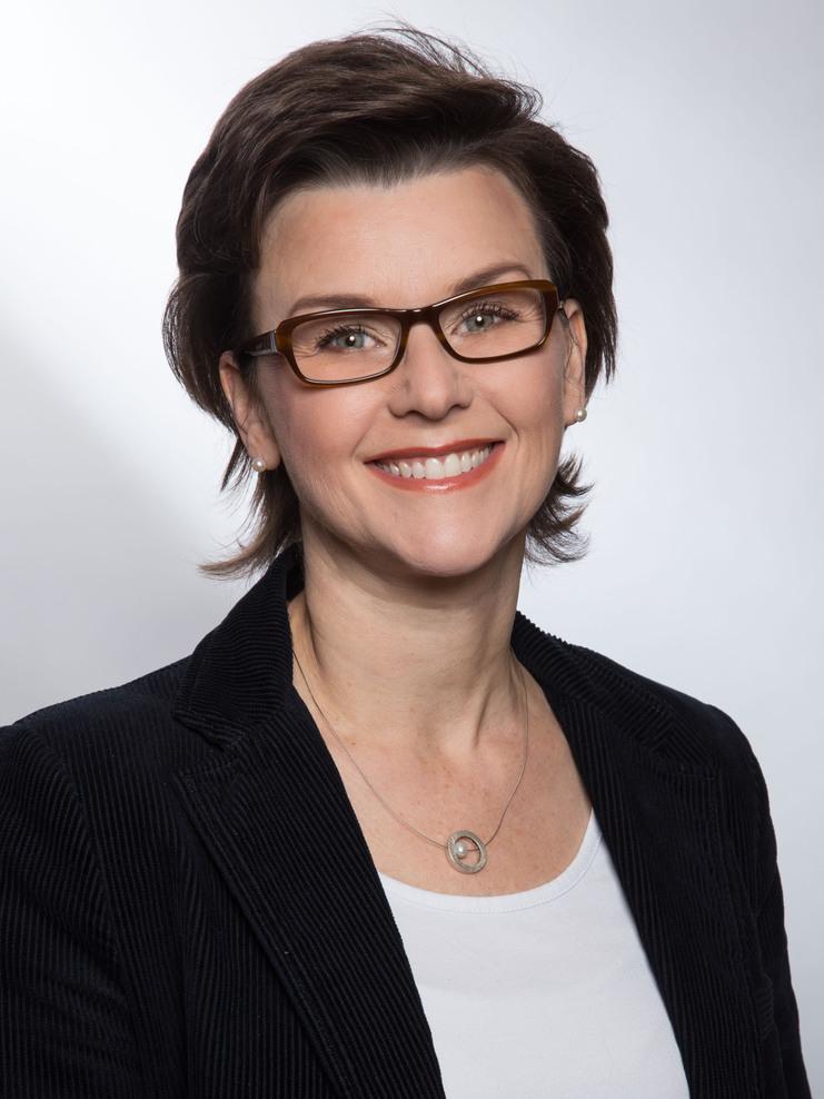 Porträt einer lächelnden Frau mit Brille, kurzen Haaren und einem schwarzen Blazer vor neutralem Hintergrund.