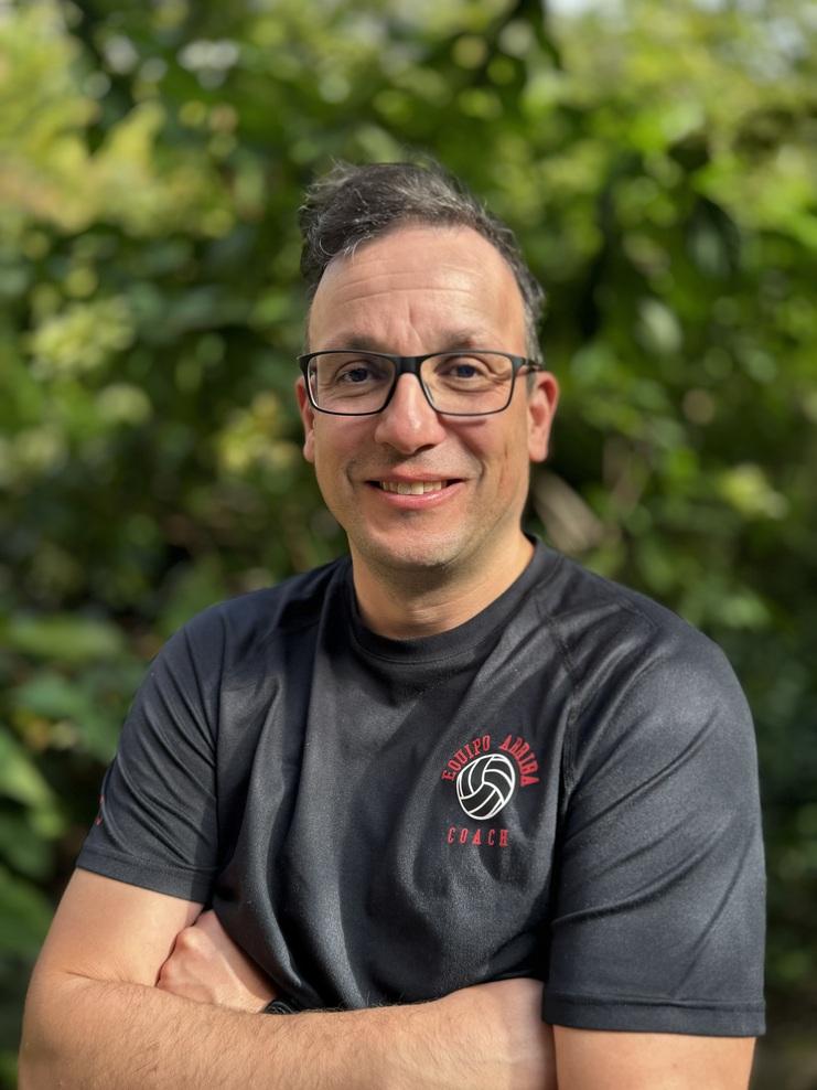 Mann mit Brille und schwarzem T-Shirt steht mit verschränkten Armen vor verschwommenem grünen Hintergrund.