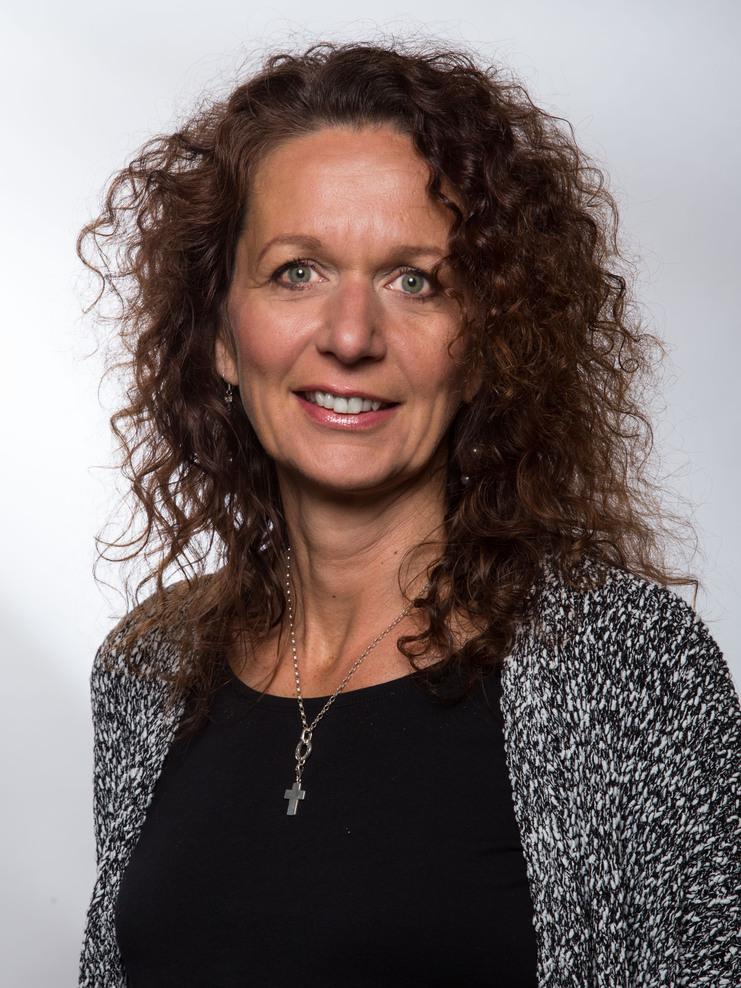 Porträt einer Frau mit lockigem, braunem Haar, schwarzem Oberteil und einer silbernen Kette mit Kreuzanhänger.