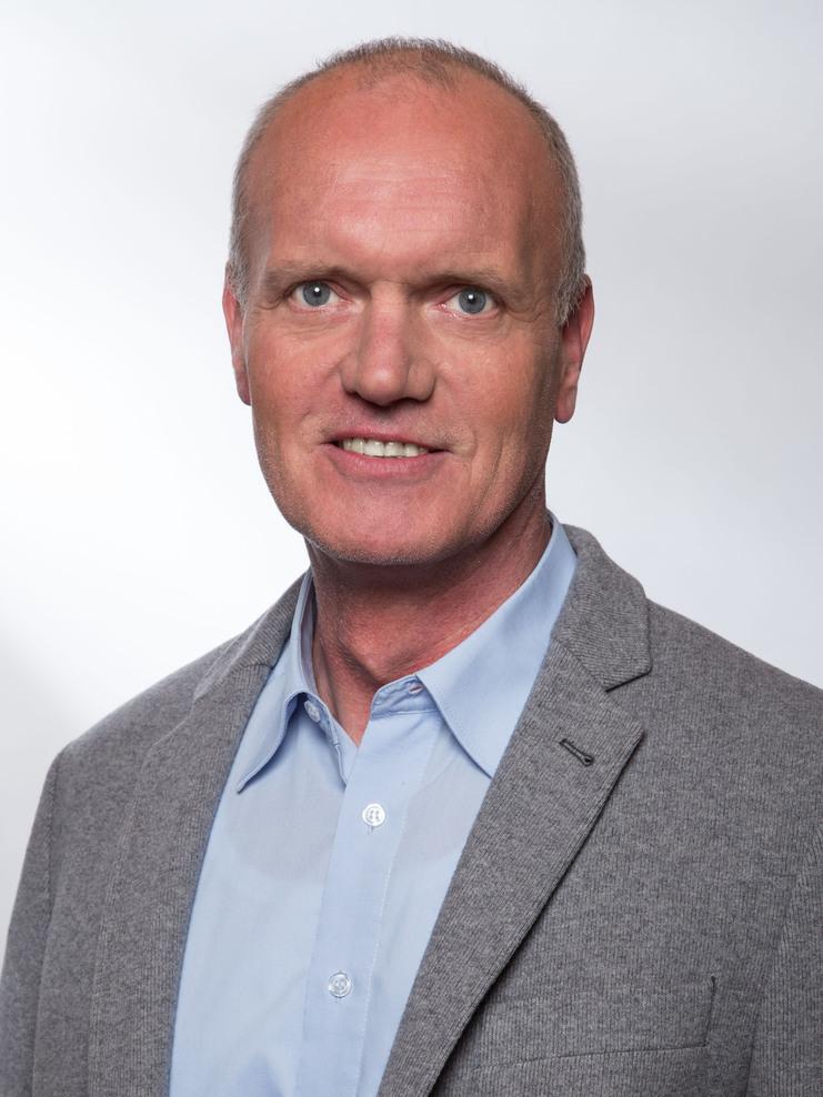 Portrait eines Mannes mit grauem Blazer und hellblauem Hemd vor einem einfarbigen Hintergrund.