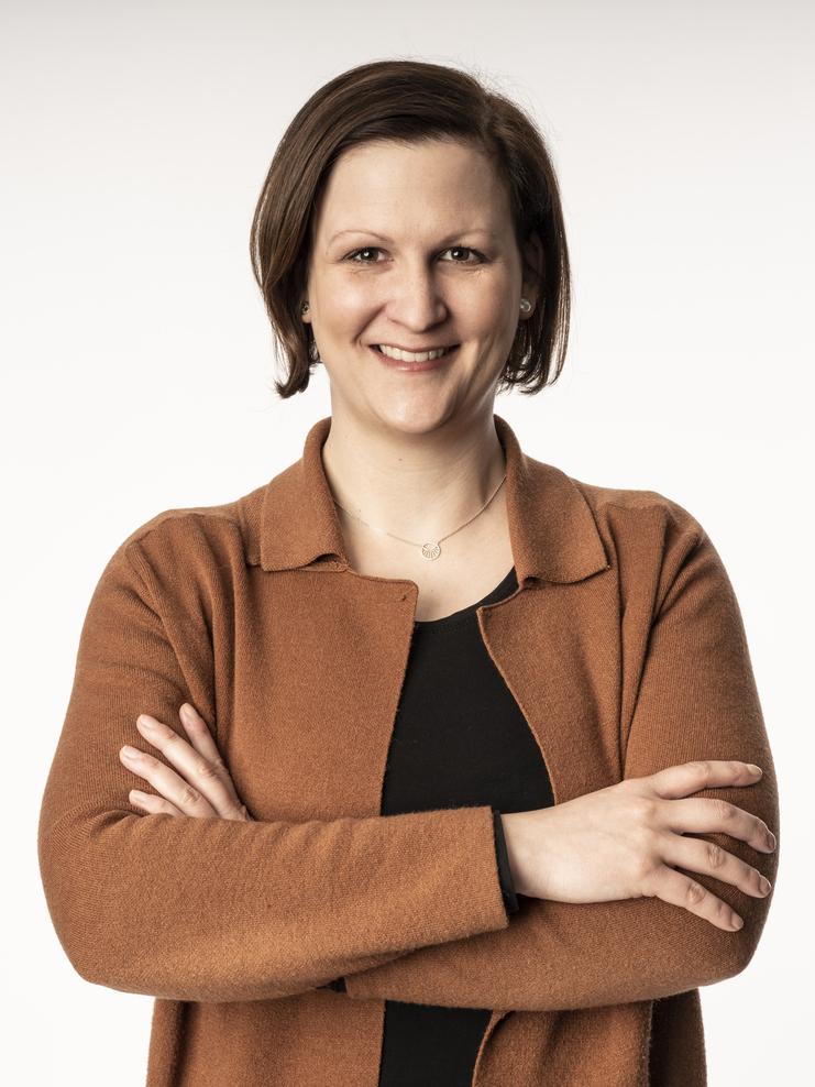 Lächelnde Person mit kurzen, braunen Haaren in einem braunen Cardigan, die mit verschränkten Armen vor einem hellen Hintergrund steht.