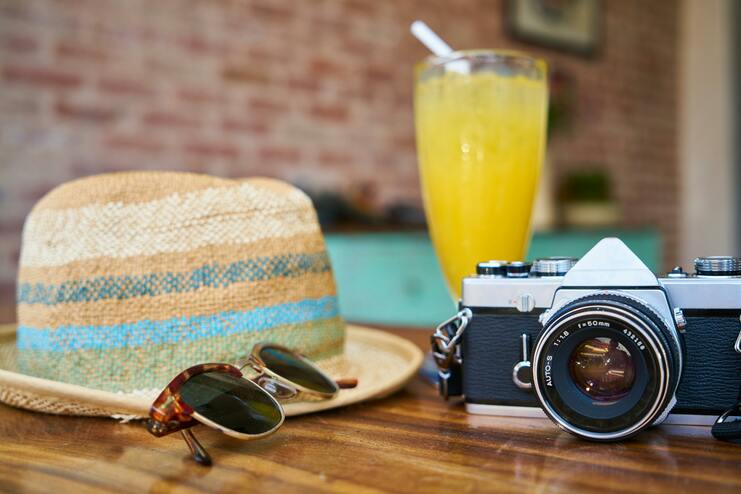 Nahaufnahme einer Kamera, Sonnenbrille und Strohut neben einem Glas mit gelbem Getr&auml;nk auf einem Holztisch.