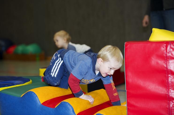 Ein Kleinkind krabbelt auf bunten Schaumstoffelementen in einer Spielhalle, während ein weiteres Kind im Hintergrund spielt.