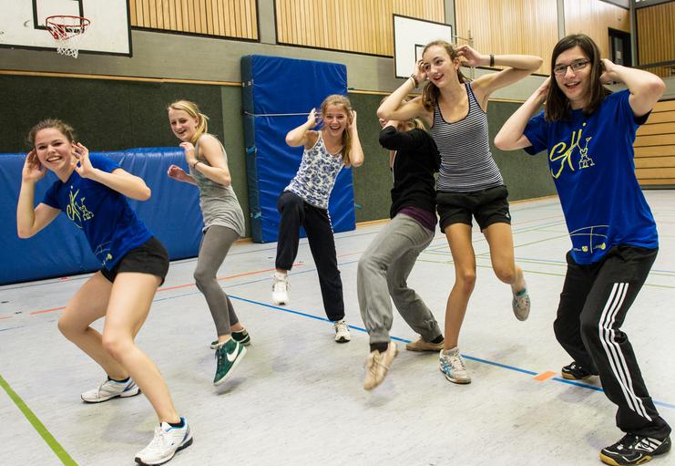 Sechs Jugendliche tanzen in einer Turnhalle, lachen und haben eine fröhliche Stimmung. Ein Basketballkorb ist im Hintergrund.