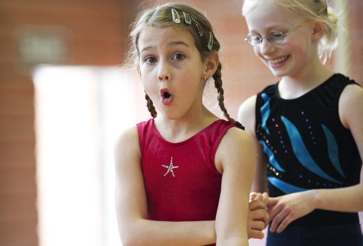 Zwei Mädchen in Gymnastikanzügen, eines überrascht und das andere lächelnd, in einer Turnhalle.