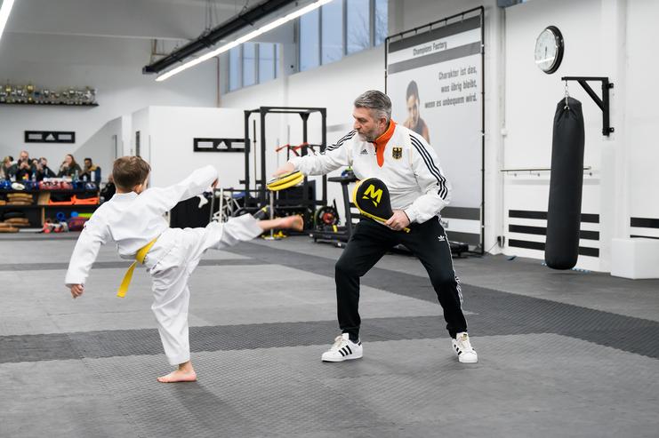 Ein Junge in Karateanzug mit gelbem Gürtel hebt das Bein zum Kick, während ein Trainer mit Pads vorbereitet.