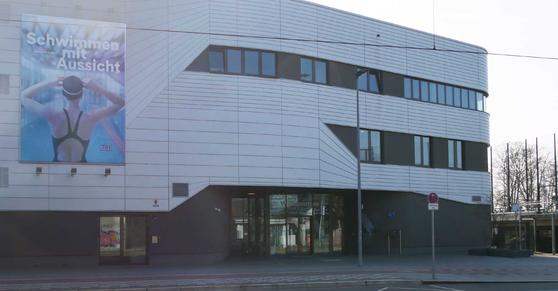 Fassade eines modernen Gebäudes mit einem großen Werbeplakat zum Schwimmen und einem Eingangsbereich.