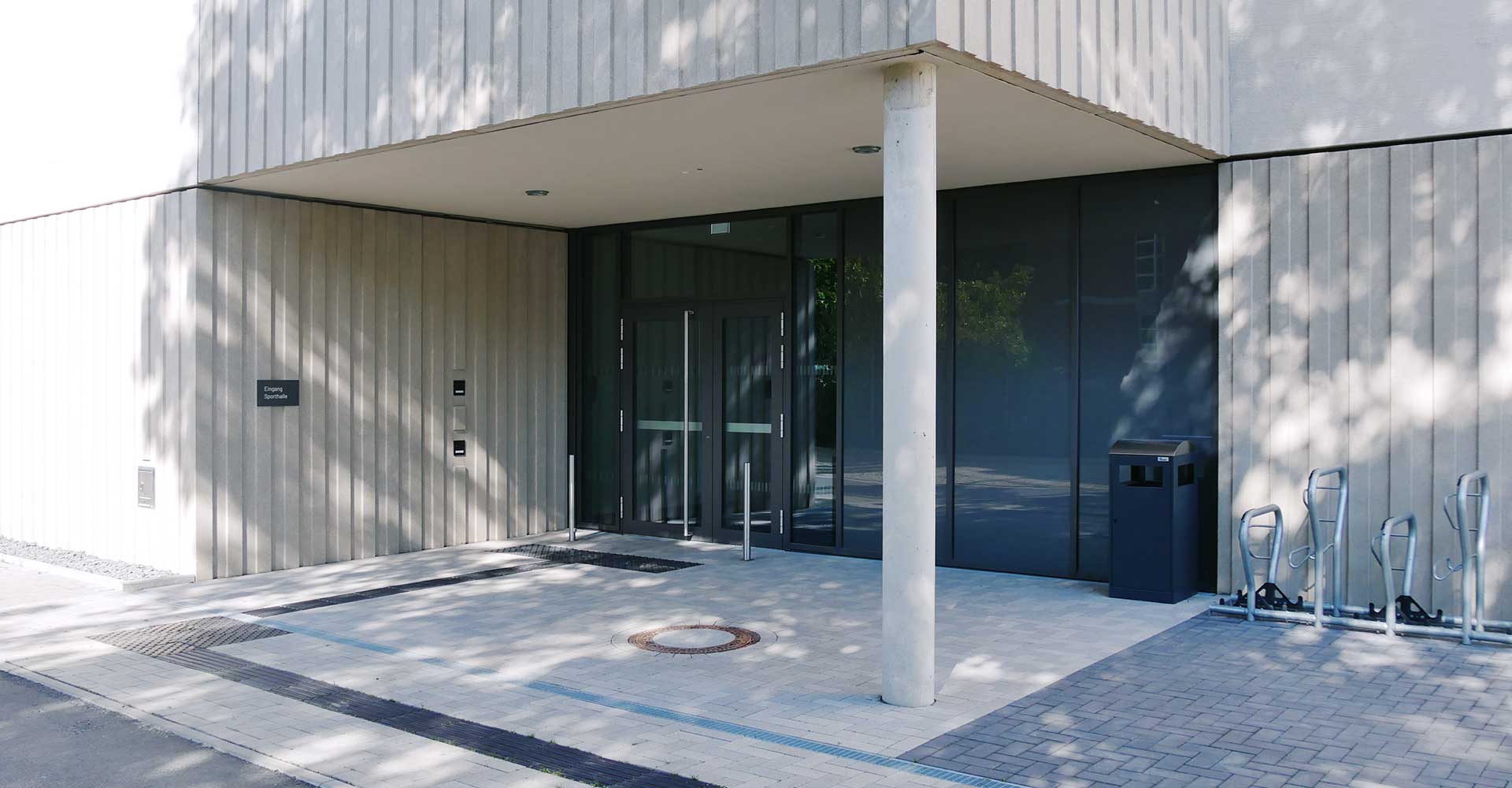 Eingang zu einem modernen Gebäude mit Glasportal, Abfallbehälter und Fahrradabstellplätzen auf dem Pflaster.